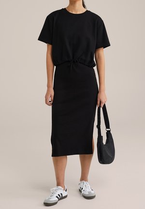 Kobieta w czarnej, krótkorękawowej bluzce z paskiem w talii i czarnej spódnicy midi z bocznym rozcięciem, białe trampki, trzymająca czarną torebkę na ramię.
