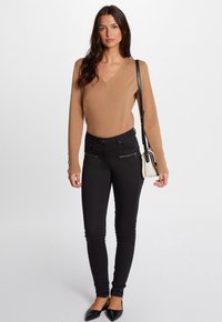 Bruine langemouwen trui met een V-hals, gecombineerd met zwarte skinny jeans met ritszakken en zwarte ballerina's. Inclusief zwarte schoudertas.