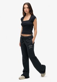 Vrouw staat rechtop, gekleed in een zwart top met korte mouwen en kantafwerking, zwarte sweatpants met trekkoord en de tekst "Superdry Athletics", en beige sneakers.