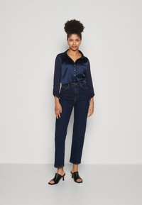 Navy Satinbluse mit Knopfdetails, kombiniert mit hochgeschnittenen, dunklen Jeans und schwarzen offenen High Heels. Glatte Texturen und maßgeschneiderte Passform.