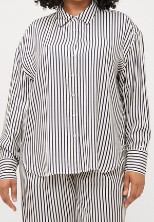 Kvinde iført løstsiddende hvid skjorte og bukser med smalle lodrette sorte striber, stående med armene afslappet ved siderne.