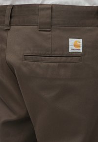 Καφέ παντελόνι από ανθεκτικό ύφασμα, με πίσω τσέπη που έχει ετικέτα με το λογότυπο Carhartt. Η υφή είναι λεία, με σχέδιο ίσιας γραμμής στα πόδια.