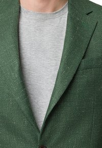 Zielona marynarka w jodełkę o teksturowanym wykończeniu, z klapami i jednolitym zapięciem na guzik, założona na szary t-shirt.