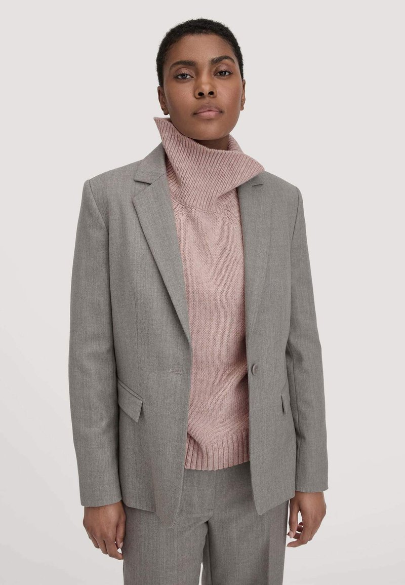 Grijze blazer over een roze geribbelde coltrui, met een dubbele knoopsluiting, gelapteloten en een op maat gesneden pasvorm.