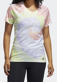 Femme portant un t-shirt tie-dye pastel à manches courtes avec le logo Adidas à l'ourlet, assorti à des leggings noirs sur fond uni.