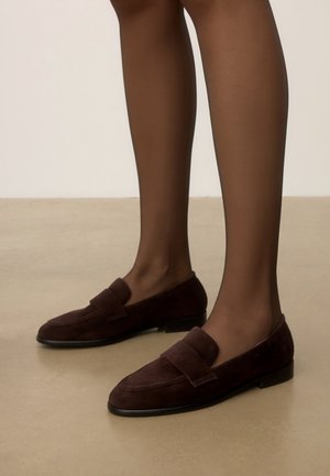 Mocassins en daim marron foncé portés pieds nus, montrés du milieu du mollet vers le bas sur un sol beige avec un fond uni.