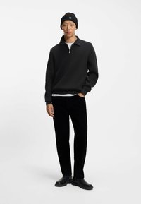 Maglione zip-up nero in tessuto testurizzato, abbinato a pantaloni in velluto a coste neri e stivali neri. La modella indossa un berretto abbinato nero.