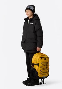 Barn som har på seg en svart vinterjakke, svarte bukser, svarte støvler og en svart lue, holder en stor gul ryggsekk i håndtaket.