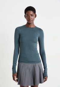 Haut à manches longues ajusté en tissu teal avec une texture lisse ; il présente un col rond et des trous pour les pouces. Associé à une jupe plissée grise.