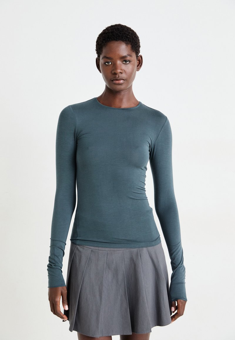 Haut à manches longues ajusté en tissu teal avec une texture lisse ; il présente un col rond et des trous pour les pouces. Associé à une jupe plissée grise.