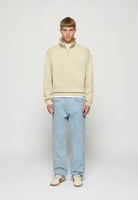 Sweatshirt bege claro com fecho éclair e gola alta, com punhos canelados, combinado com jeans de perna reta azul claro e ténis creme.