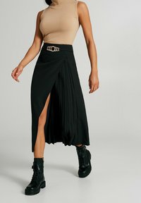 Gonna nera plissée con uno spacco alto, caratterizzata da un dettaglio cinturino in tonalità oro. Indossata con un dolcevita beige e stivali neri con lacci.