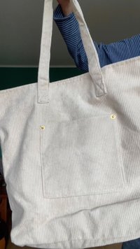 Saco de tela com riscas verticais brancas e bege claro, duas alças robustas e um bolso frontal seguro com botão de pressão dourado.