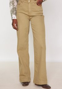 Beige Hochwasserhose aus Baumwolle mit einem hohen Bund, zwei Seitentaschen und einem Knopfverschluss. Glatte Textur, taillierte Passform.