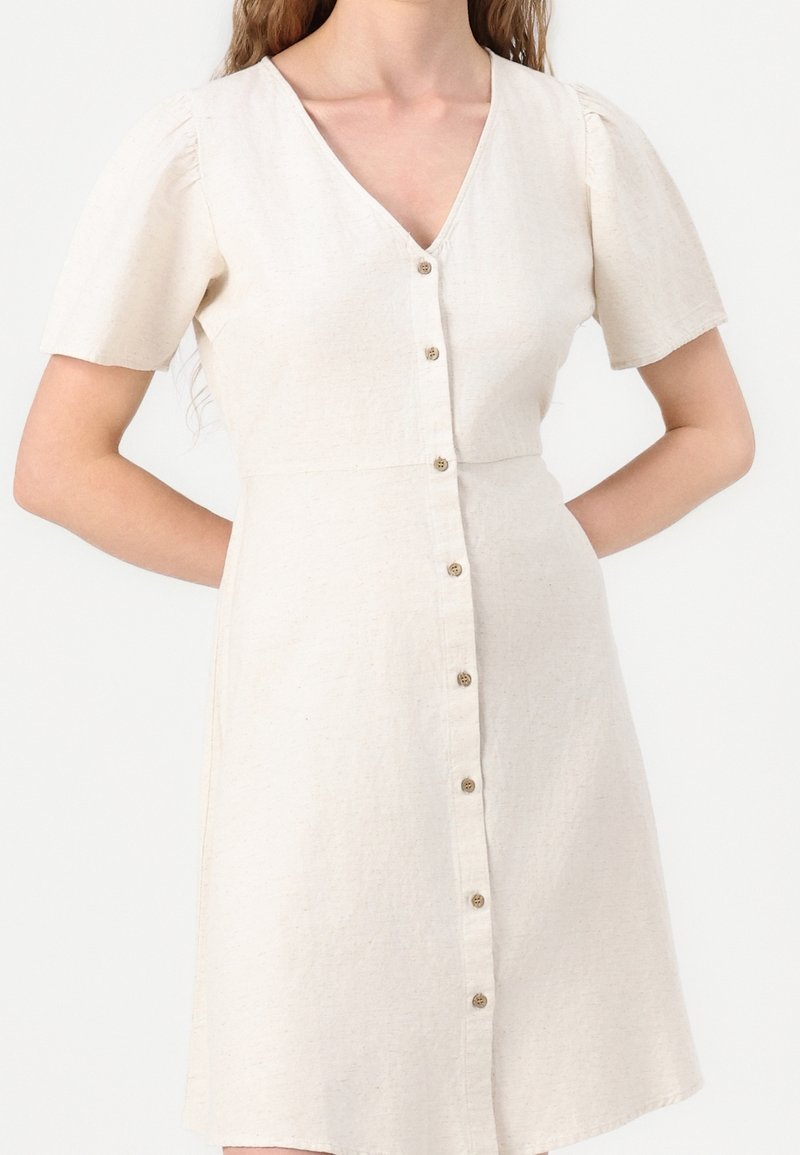 Femme portant une robe à manches courtes, beige clair, boutonnée sur le devant avec un col en V, debout avec une main sur la hanche sur un fond uni.