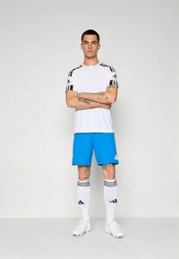 Wit sportshirt met zwarte strepen, felblauwe korte broek, witte sokken met zwarte accenten en witte voetbalschoenen.