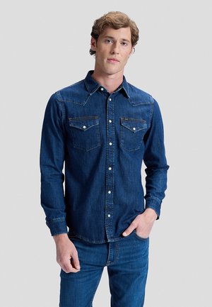 Jeune homme portant une chemise en denim bleu foncé boutonnée avec des poches à rabat et un jean assorti, debout devant un fond clair uni.