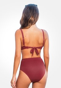 Donna con lunghi capelli che indossa un bikini bottom a vita alta color marsala e un bikini top con lacci, in piedi su uno sfondo chiaro e semplice.