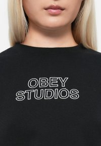 Mustaripullover, millel on julge valge brodeeritud tekst "OBEY STUDIOS". Ümar kaelus ja sile kangas.