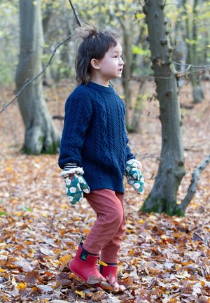 Child in a navy cable-knit sweater, pink pants, red waterproof boots, and green floral mittens, walking among autumn leaves in a forest.