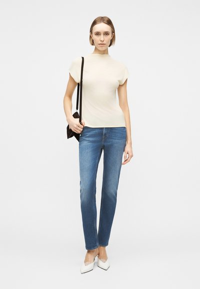 Γυναίκα που στέκεται, φοράει κρεμ κοντομάνικη μπλούζα, μπλε τζιν, λευκά μυτερά παπούτσια και κρατάει μια μικρή μαύρη τσάντα ώμου.