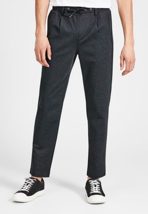 Homme portant un pantalon fuselé gris foncé avec une taille à cordon et des baskets basses noires avec des lacets et semelles blanches.