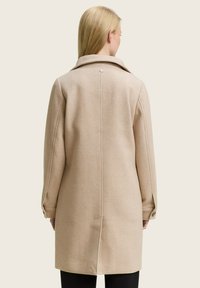 Vrouw in een beige wollen jas met een achterkantventilatie, knopen bij de manchetten en een klein rond metalen detail onder de kraag, staand tegen een effen achtergrond.