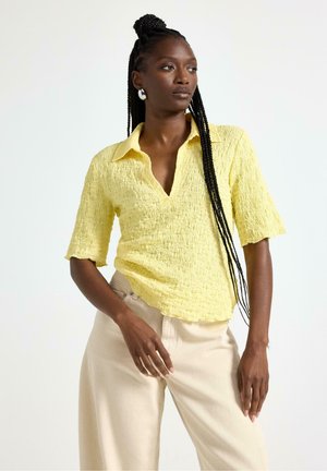 Kvinde med langt fletning hår iført tekstureret gul kortærmet skjorte og beige bukser, poserer med den ene hånd i lommen og kigger til siden.