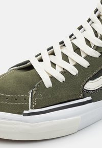 Grønn semska sneaker med hvite snører, mønstrede detaljer og svart gummisåle. Har kontrasterende sømmer og en teksturert overflate.