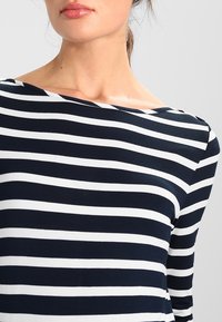 Granatowo-biały top z długim rękawem w paski z dekoltem w łódkę. Gładka bawełniana tkanina z równomiernymi poziomymi paskami. Brak widocznych elementów metalowych czy ozdób.