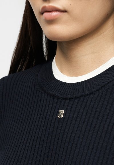 Κοντινό πλάνο ενός ατόμου που φοράει μπλε πουλόβερ με ριγέ ύφανση και ένα μικρό μεταλλικό διακοσμητικό σήμα κοντά στη λαιμόκοψη πάνω από ένα λευκό εσώρουχο.