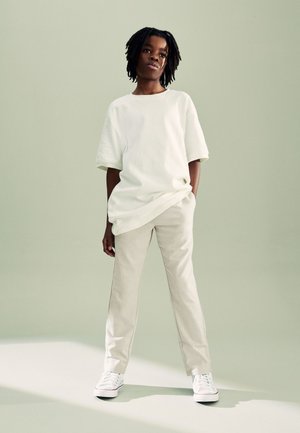 Osoba s kratkim dreadlocks stoji sa rukama u džepovima, noseći preveliku bijelu majicu, bež hlače i bijele tenisice na svijetloj pozadini.