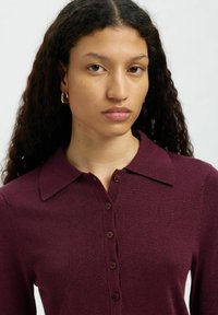 Pikkade lokkis juustega naine, kes kannab väikseid kuldseid rõngaskõrvarõngaid ja burgundi tooni nööbitavat kudumit kraega, lihtsa tausta ees.