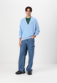 Cardigan blu chiaro con scollo a V, chiusura con bottoni e orlo a coste; indossato sopra una camicia verde. Abbinato a pantaloni in denim larghi.