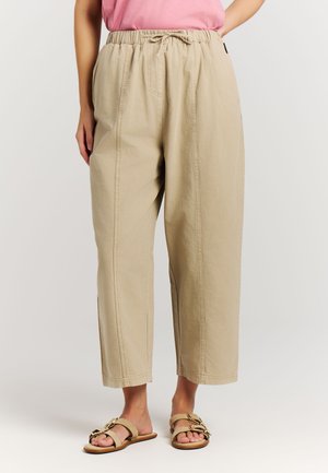 Vrouw die beige, wijd uitlopende, cropped broek draagt met een elastische tailleband met trekkoord en beige sandalen met gespen.