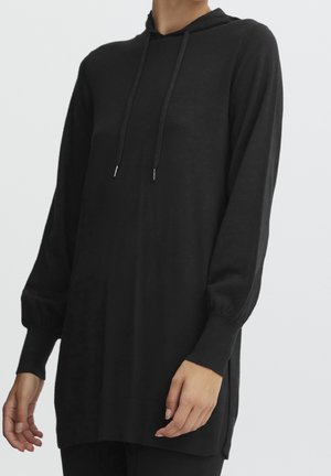 Femme portant un long sweat à capuche en maille noire avec des poignets côtelés et des cordons de serrage, présenté du cou au milieu des cuisses sur un fond uni.