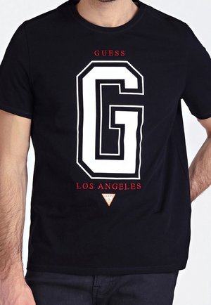T-shirt noir avec un grand "G" blanc en contour, texte rouge "GUESS" au-dessus et "LOS ANGELES" en dessous, porté par une personne les mains sur les côtés.