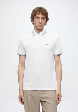 Jeune homme aux cheveux châtain clair portant un polo blanc à manches courtes avec le logo "BOSS" et un pantalon beige sur un fond uni.