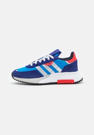 Blauer und weißer Sportschuh mit roten Akzenten, weißen Schnürsenkeln und einer dicken weißen Sohle mit schwarzem und rotem Profil.