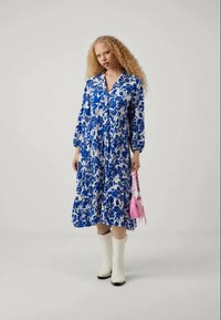 Bloemdruk jurk in blauw en wit met lange mouwen en een gelaagde zoom. Gepaarde met witte laarzen en een kleine roze handtas.