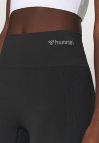 Close-up van een persoon die zwarte naadloze leggings met hoge taille draagt en een witte sporttop met het hummel-logo op de tailleband.