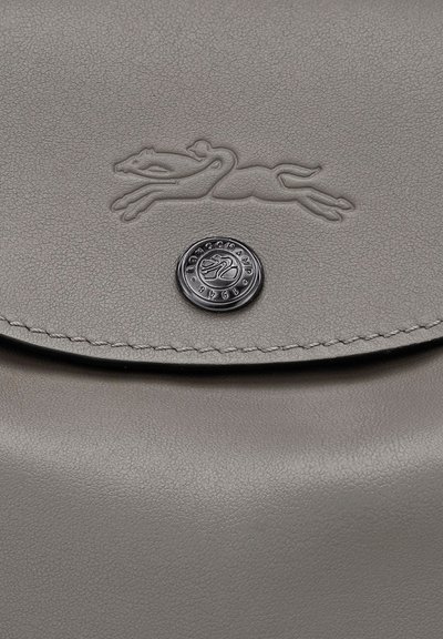 Graue Lederhandtasche mit eingeprägtem Laufgreyhound-Logo und einem runden Metallknopf, der mit dem Markennamen und dem Jahr 1948 graviert ist.