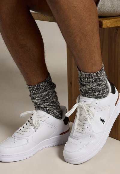 Beine mit dunkler Haut, die graue gestrickte Socken und weiße Polo Ralph Lauren Sneaker tragen, sitzen auf einer Holzbank vor einem hellen Hintergrund.