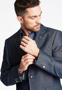 Dunkelblaues Denim-Blazer mit Reverskragen, einreihiger Knopfschluss und geknöpften Manschetten mit dekorativer Naht. Darunter ein gestreiftes Hemd.