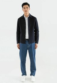 Svart teksturert jakke med knapper over en hvit stripete skjorte, sammen med blå denimjeans og hvite sneakers. Foran.