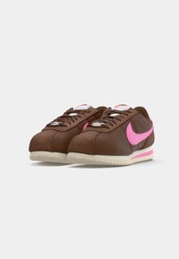 Pár hnědých tenisek Nike s růžovými swooshi, bílými středními podrážkami s růžovými pruhy a béžovými texturovanými podrážkami, zobrazenými pod mírným úhlem.