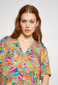 Blusa floral multicolorida com decote em V, mangas curtas, padrões vibrantes de rosa, amarelo e azul em um tecido texturizado.