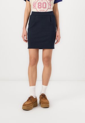 Kobieta ubrana w granatową mini spódniczkę z przednimi kieszeniami, białe skarpetki, brązowe buty na platformie i casualowy t-shirt z grafiką włożony do środka.