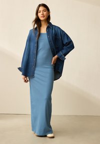 Denim overhemd, oversized pasvorm met opgestikte zak. Lichtblauwe geribbelde maxi-jurk. Witte schoenen. Neutrale achtergrond en minimalistische setting.