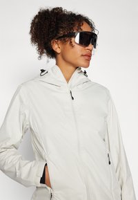 Lättvit zip-up jacka med huva, med svarta toggles och fickor med dragkedja. Kommer med stora svarta solglasögon.
